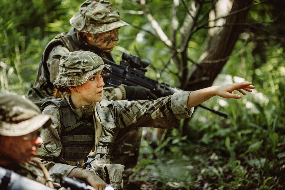 Three soldiers in camouflage uniforms in a wooded area, with one person pointing ahead