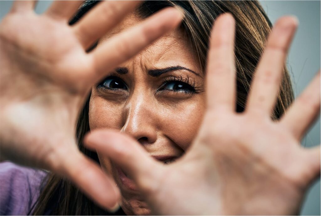 Frightened woman raising hands to shield herself, face centered between outstretched palms, symbolizing domestic violence and fear.