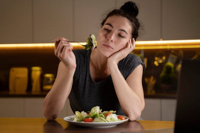Woman sitting at a table holding a fork with salad, resting her head on her hand in a kitchen setting thinking about anxiety and nutrition
