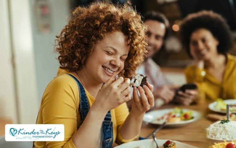 Woman smiling while holding a small dessert at a dining table with two people in the background